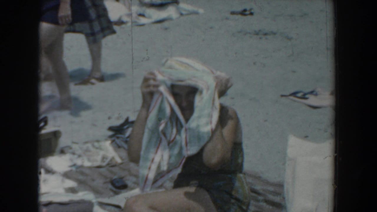 1958 Yorktown beach home movie frame with a seated beachgoer drying her head under a striped towel, sandals and legs nearby.