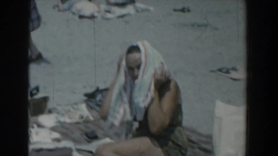 In the Yorktown, Virginia 1958 beach footage, a woman in a swimsuit sits on the sand and dries her hair with a striped towel.