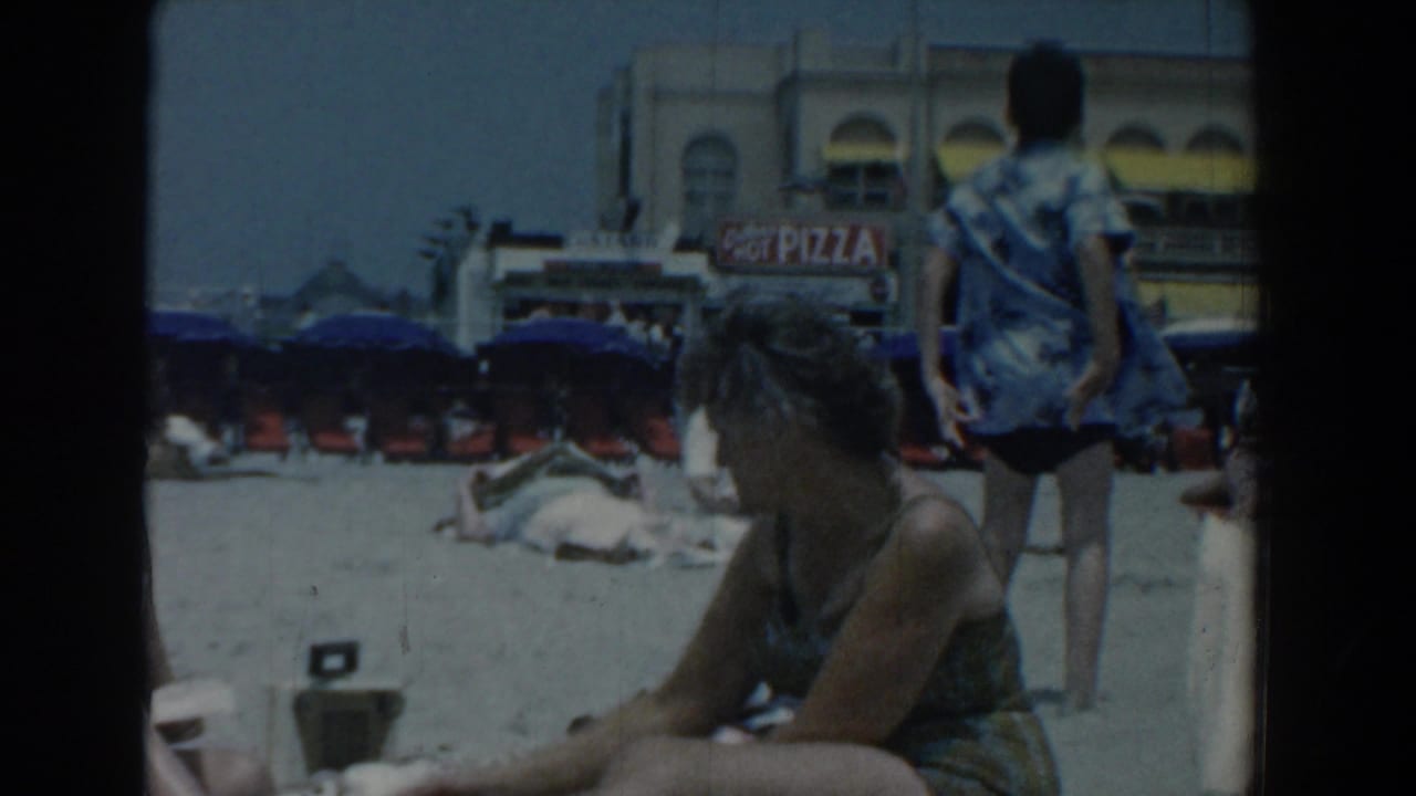 Beachgoers relax on the Yorktown sand in 1958, with rows of umbrellas, beachfront buildings, and a visible pizza sign behind them.