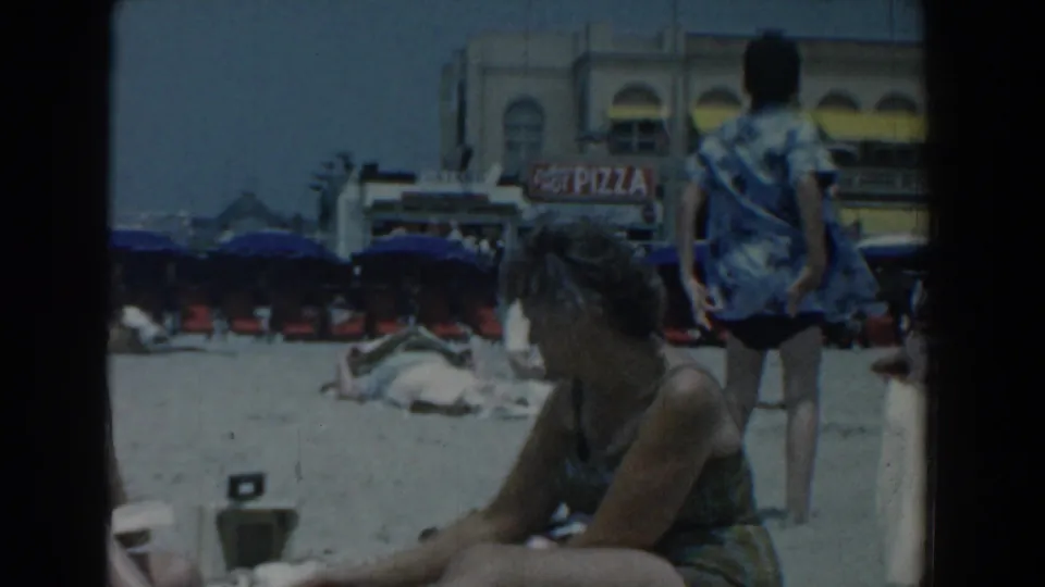 Beachgoers relax on the Yorktown sand in 1958, with rows of umbrellas, beachfront buildings, and a visible pizza sign behind them.