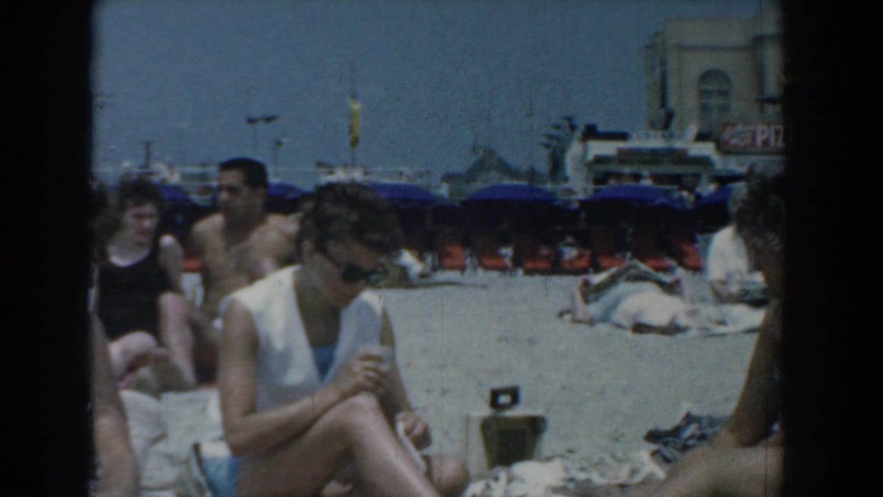 A candid 1958 Yorktown beach view shows several adults seated on towels, with blue umbrellas, sand, and beachfront structures in the background.