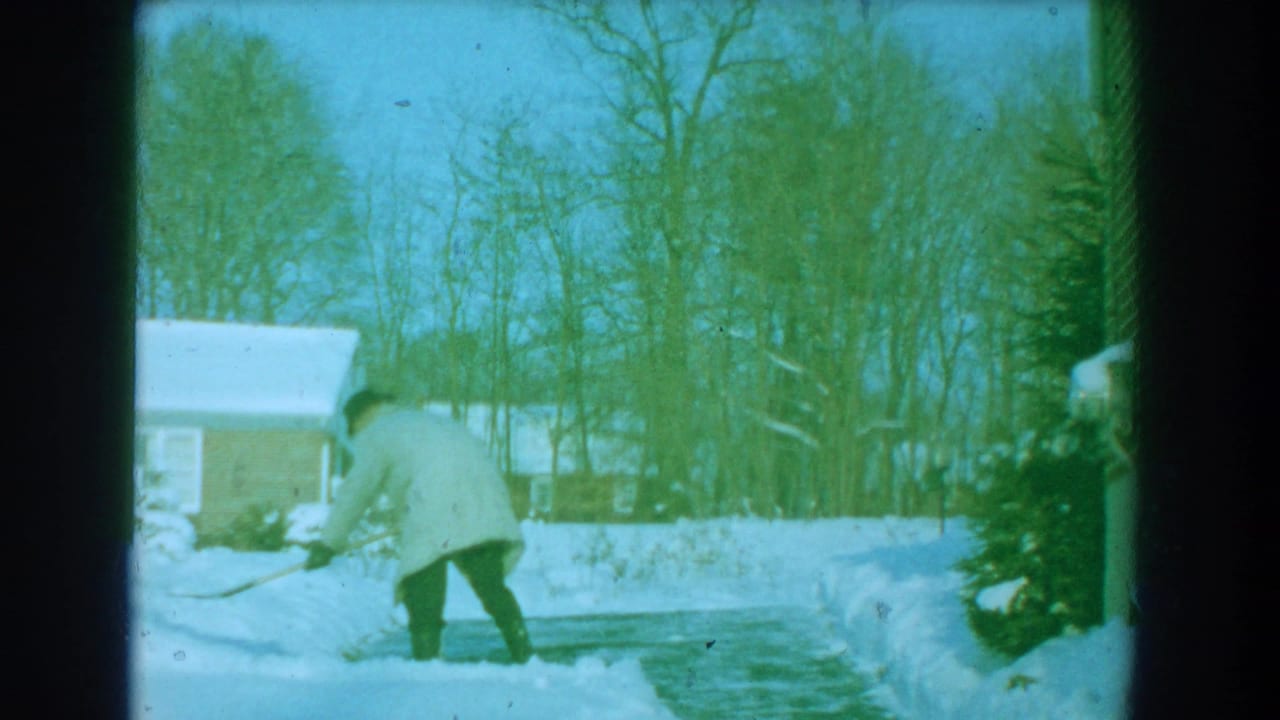 1959 Florida home-movie frame showing a man in a light coat shoveling snow near a driveway, with a house and bare trees in the background.