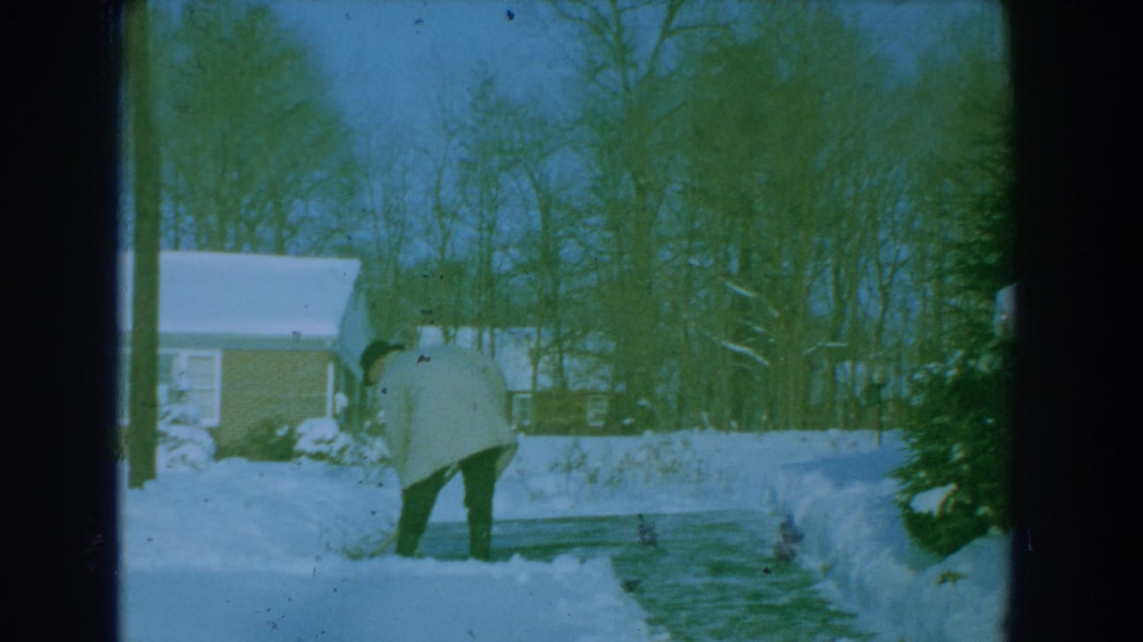 Man in a light coat shovels snow on a residential driveway near a brick house.