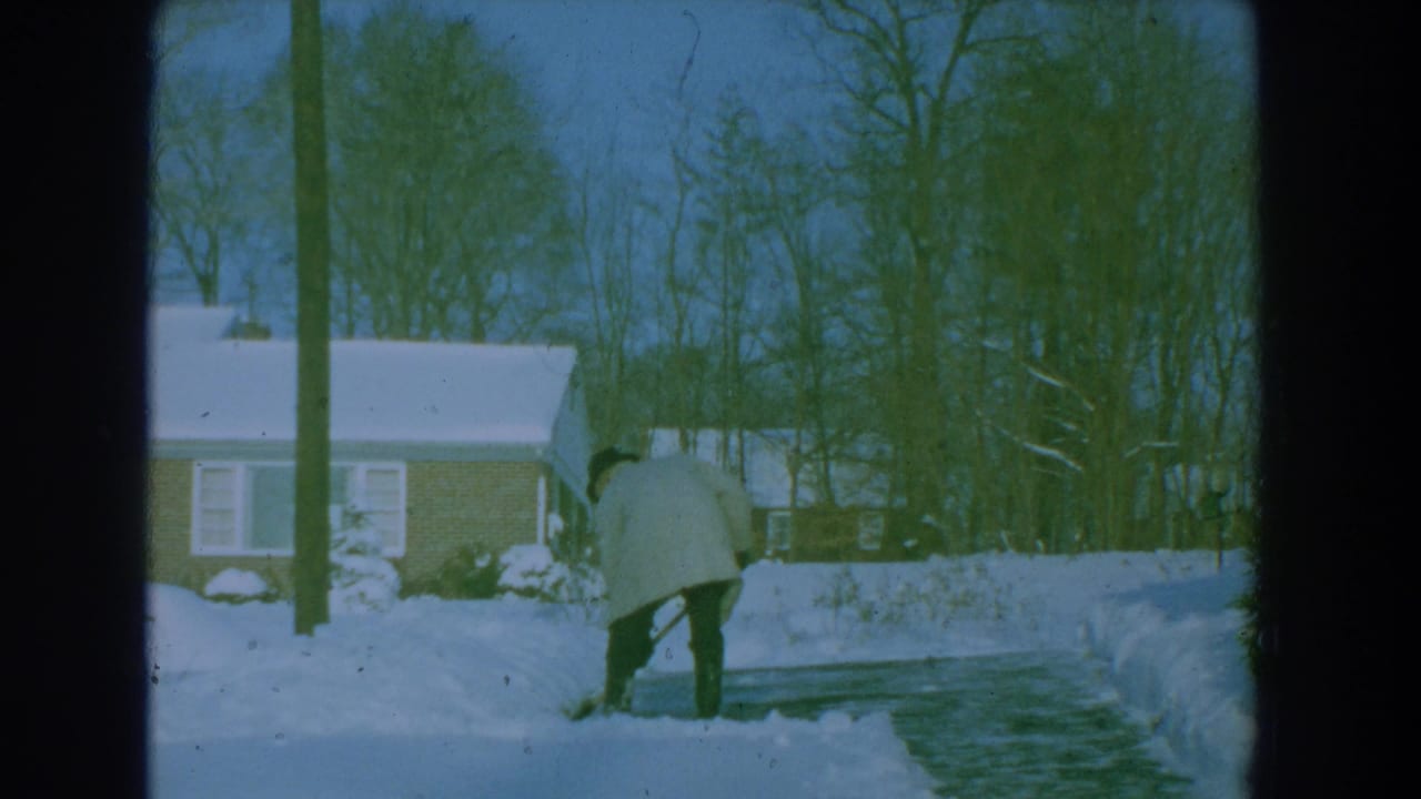 Man bends with a shovel beside a cleared path through snow in front of a house.