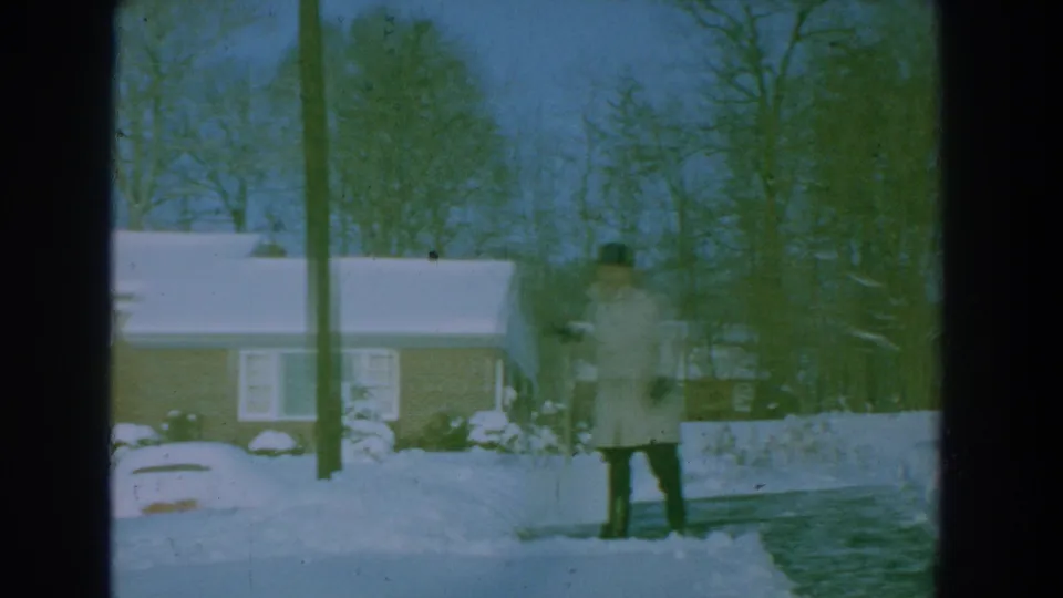 Motion-blurred 1959 8mm frame shows the man pausing or moving with a shovel in a snowy residential driveway scene.