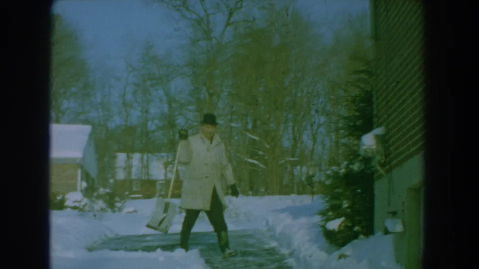 The man walks with a snow shovel beside a cleared driveway, with snowbanks, a house, and winter trees visible in the frame.