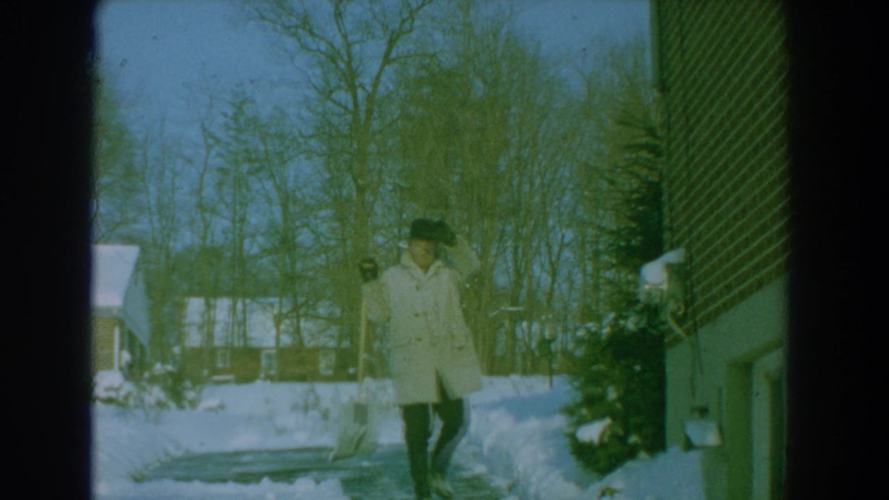 1959 8mm frame showing a man in winter clothing near a snow-covered driveway and brick house, with bare trees in the background.