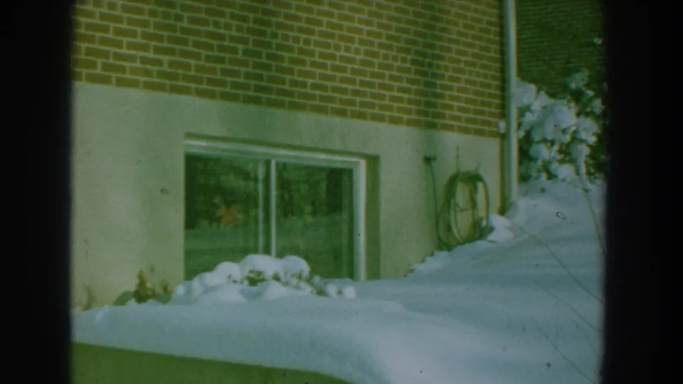 1959 8mm frame of a snowy residential exterior with brick wall, low window, coiled hose, shrubs, and snowbanks.