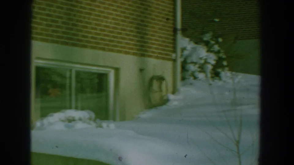 1959 8mm frame showing snow piled against a brick residential wall, with a low window, hose, shrubs, and bare branches.