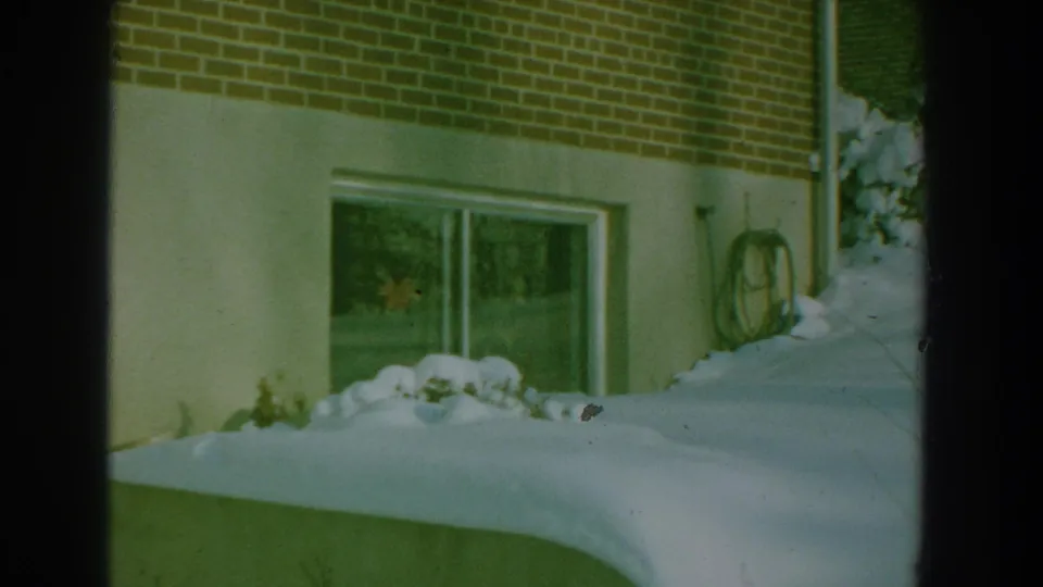 1959 8mm frame of a snow-covered residential side yard with a brick wall, low window, coiled hose, shrubs, and deep snow.