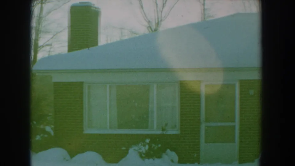 1959 Florida 8mm frame of a brick house front with snow on the roof, a chimney, large window, entry door, and shrubs.