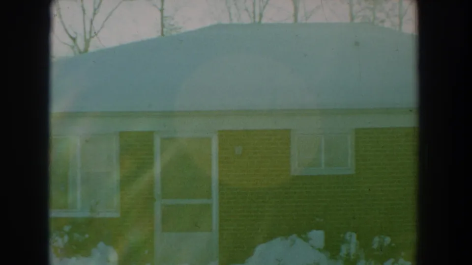 1959 Florida 8mm frame showing the house roof heavily covered with snow above a brick wall, door, window, shrubs, and bare trees.