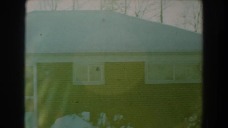 1959 Florida 8mm frame focused on a snow-covered roofline above a brick wall with small windows, shrubs, and winter trees behind.