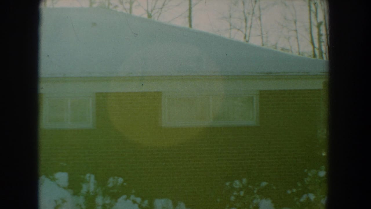 1959 Florida home-movie frame showing a snow-covered brick house, bare trees, and dark window-frame edges in the foreground.