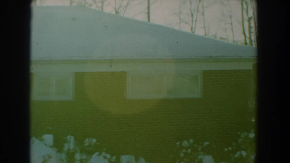1959 Florida home-movie frame showing a snow-covered brick house, bare trees, and dark window-frame edges in the foreground.