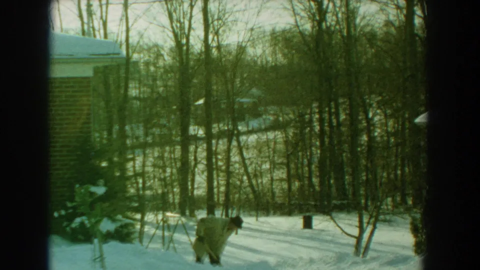 The man continues shoveling snow along a path beside a brick house, with bare trees and long shadows visible.
