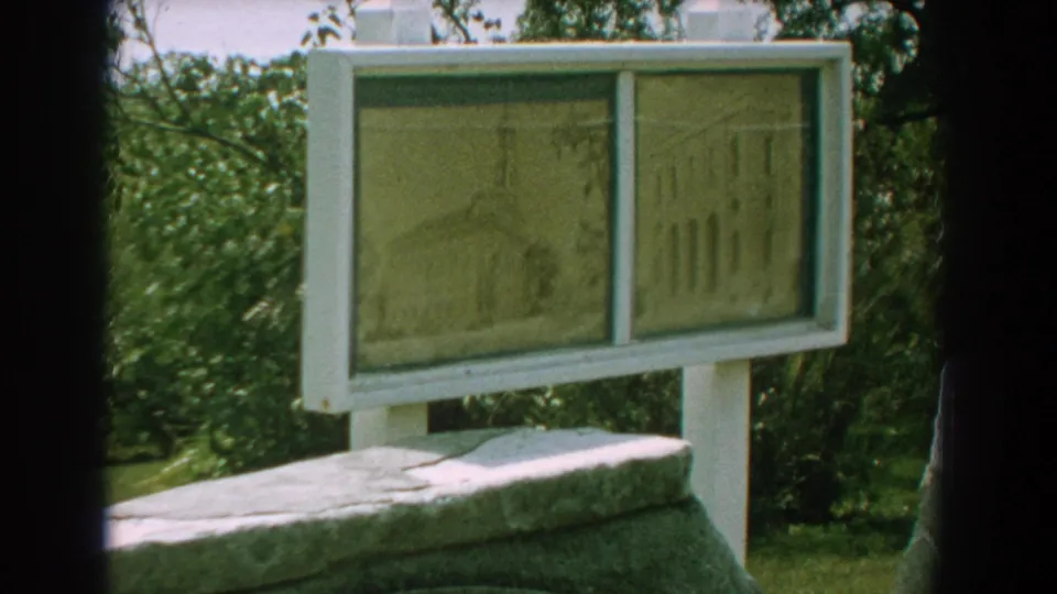 Florida 1959 8mm frame showing a white-framed interpretive sign with two sepia building illustrations above a stone marker.