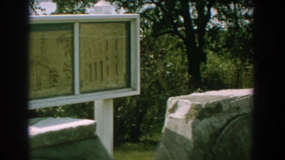 1959 Florida 8mm frame showing outdoor historical sign panels near grey stonework, green trees, and dark film edges.
