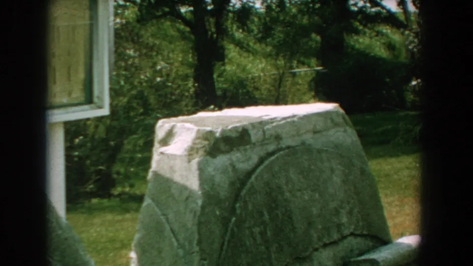 1959 Florida home-movie frame with a rough grey stone monument, partial sign panel, grass, trees, and heavy film-frame shadow.