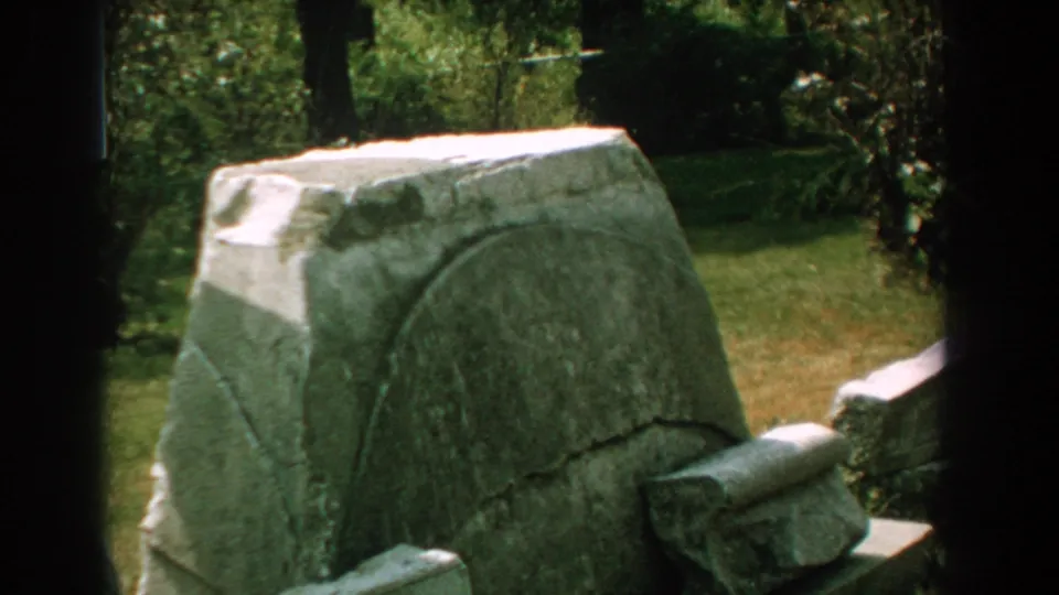1959 Florida 8mm frame focused on a weathered grey stone monument with carved curved lines, lawn, trees, and partial stonework nearby.