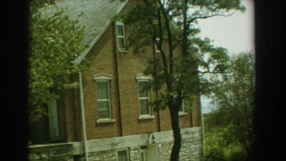 Tilted 1959 Florida 8mm view of a brick house with white-trimmed windows, roof edge, trees, and stone foundation.