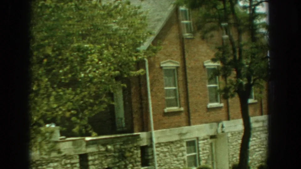 1959 Florida 8mm frame with leafy foreground growth partly covering a brick house, white-trimmed windows, and stone foundation.