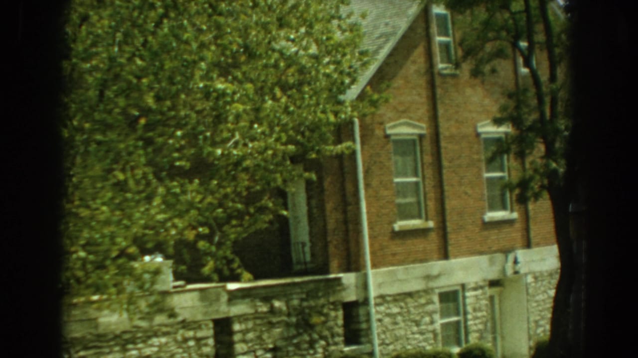 1959 Florida 8mm frame showing a brick residential house partly hidden by green trees and a low stone wall.