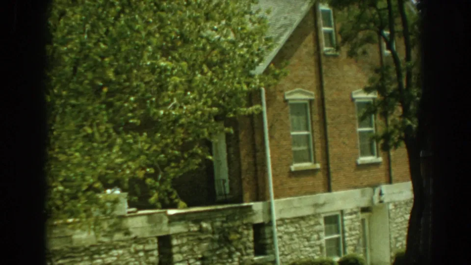1959 Florida 8mm frame showing a brick residential house partly hidden by green trees and a low stone wall.