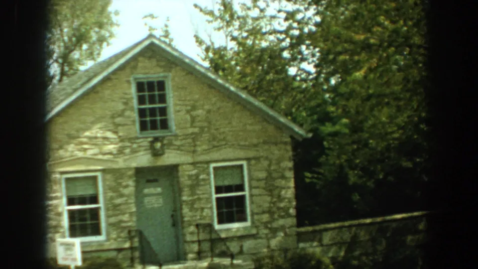 1959 Florida 8mm frame showing the front of a tan stone building with a green door, windows, railings and a small sign near the entrance.