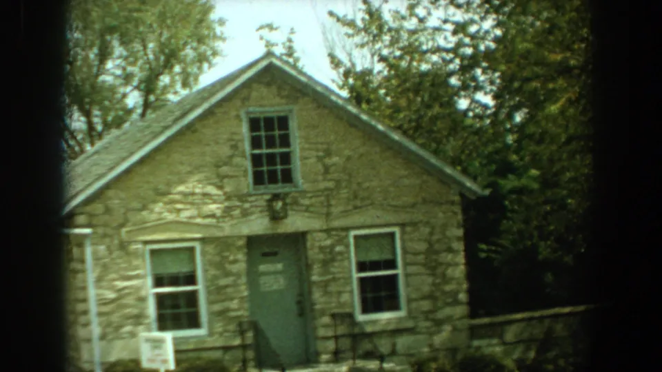1959 Florida frame with a tan stone building, green entrance door, white-trimmed windows, trees and a low stone wall.