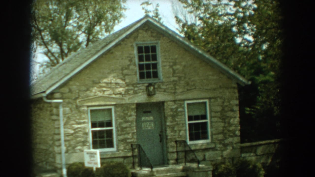 Stone cottage with gabled roof, front door, windows, trees, and a small yard sign.