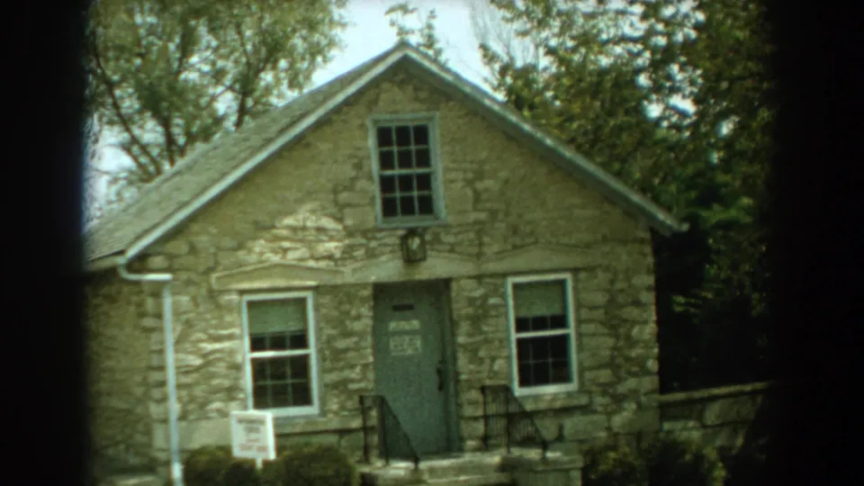 1959 Florida 8mm frame showing a rustic stone house exterior with a gable, front steps, windows, trees, and a posted sign in the yard.