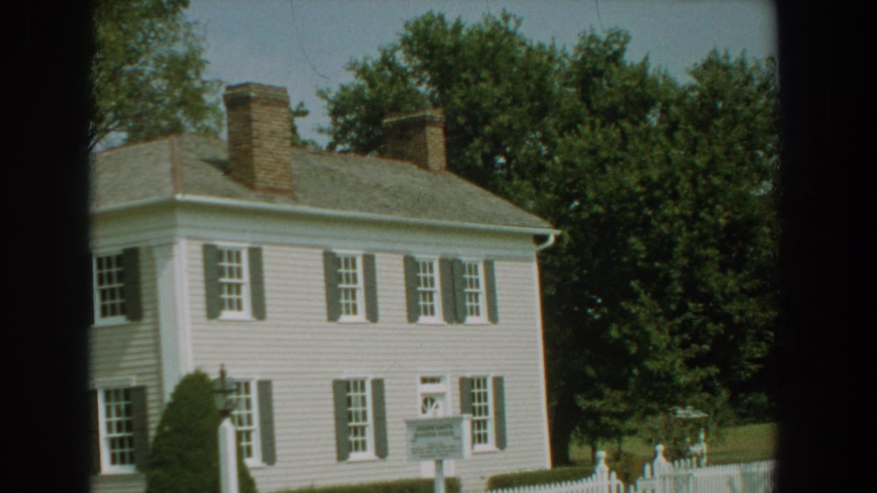 White clapboard house with dark shutters, two chimneys, trees, and a yard sign.