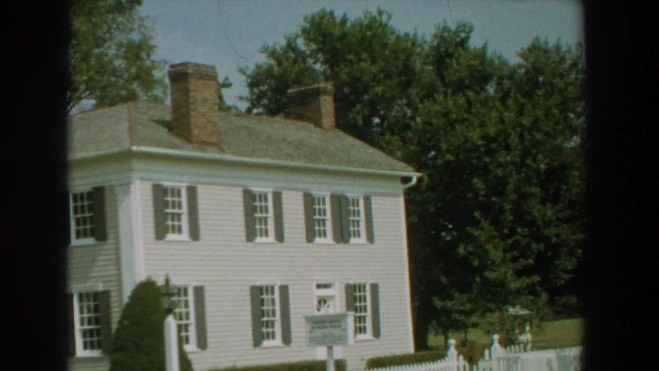 1959 Florida 8mm frame of a white clapboard historic house with dark shutters, brick chimneys, trees, shrubs, and a sign near the lawn.