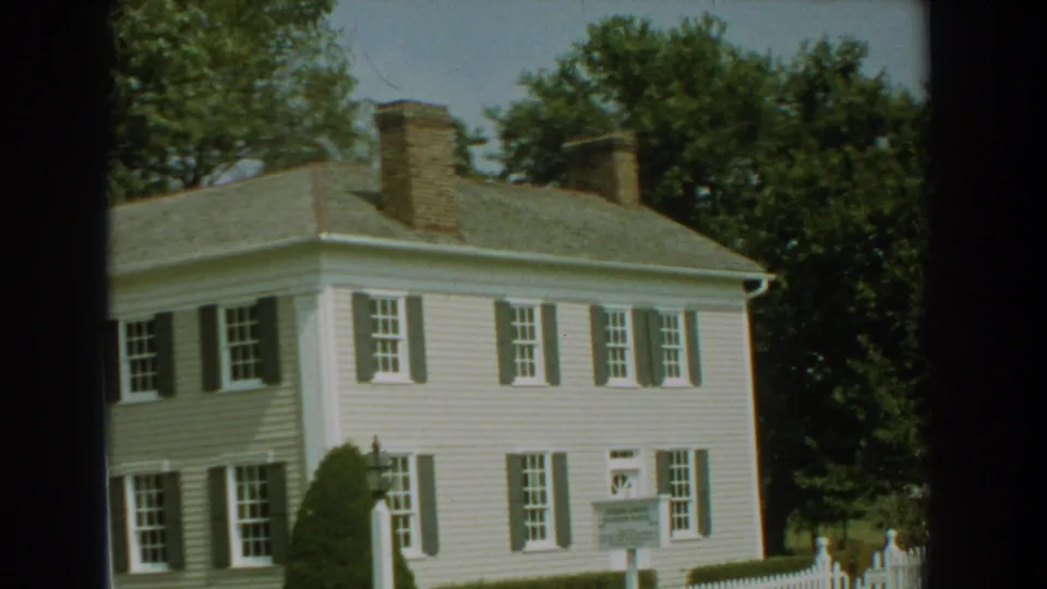 1959 Florida 8mm frame showing the white clapboard house from an angled view, with shuttered windows, chimneys, trees, shrubs, and a lawn sign.