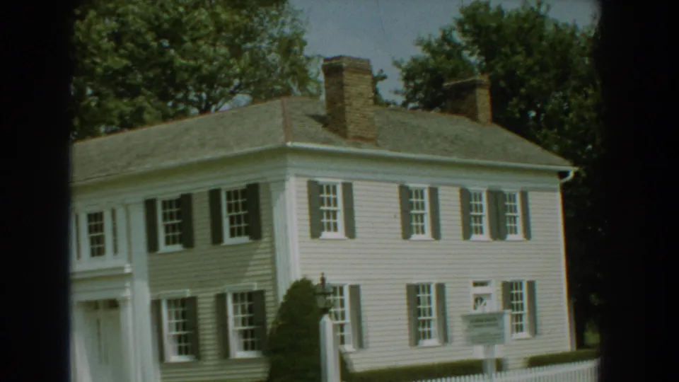 1959 Florida 8mm frame of a two-story white house with dark shutters, front entry columns, brick chimneys, trees, and a sign by the fence.