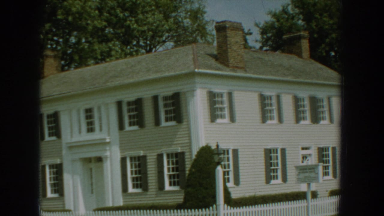 1959 Florida 8mm frame showing a white colonial-style house with dark shutters, columns and a picket fence.