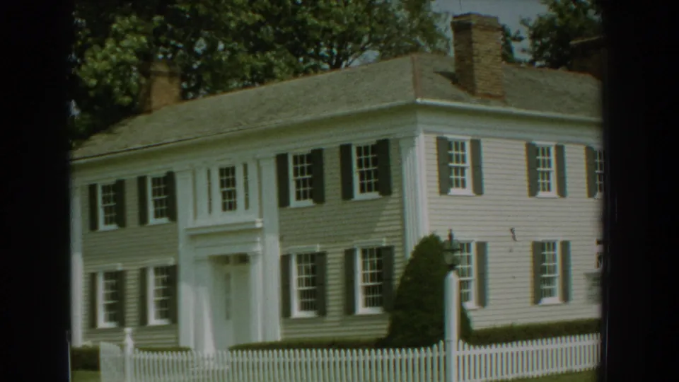 1959 Florida 8mm frame with an angled view of a white clapboard house, picket fence and trees.