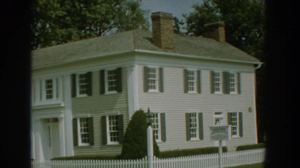 1959 Florida frame showing the house exterior, picket fence, marker sign and shaded trees on the lot.