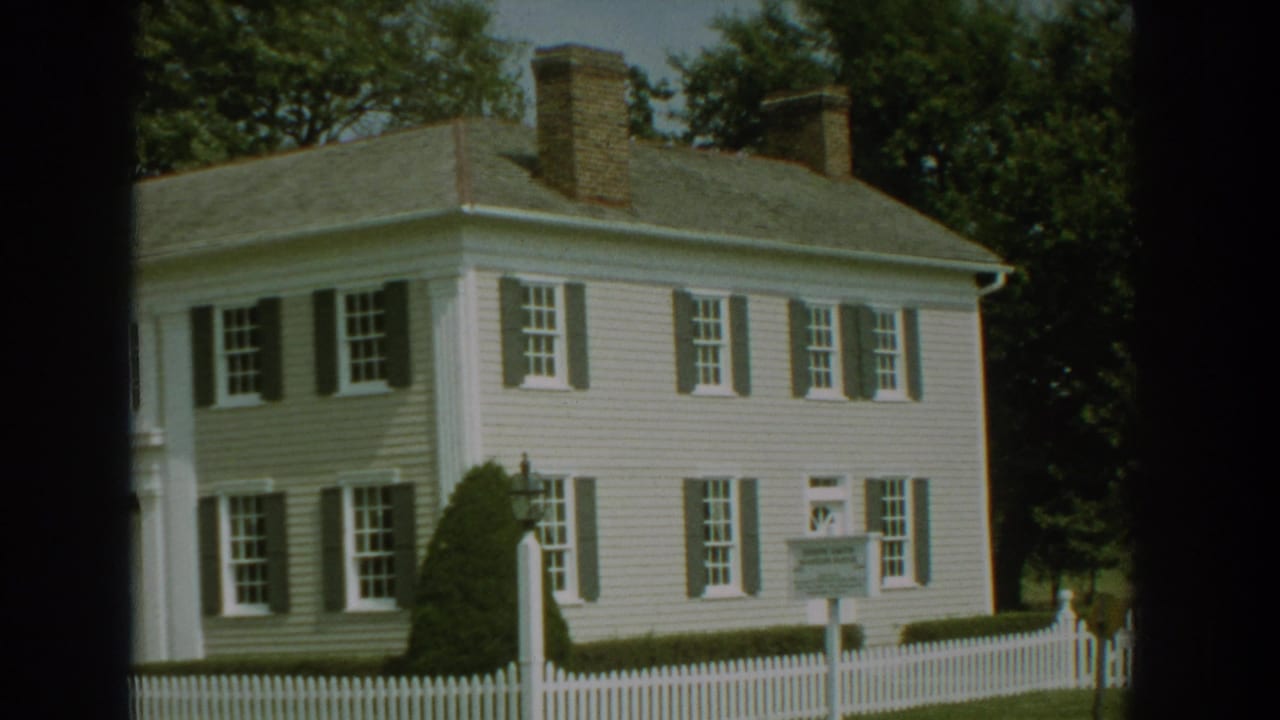 1959 Florida 8mm frame showing a white two-story house with dark shutters, brick chimneys, hedges, trees, and a picket fence.