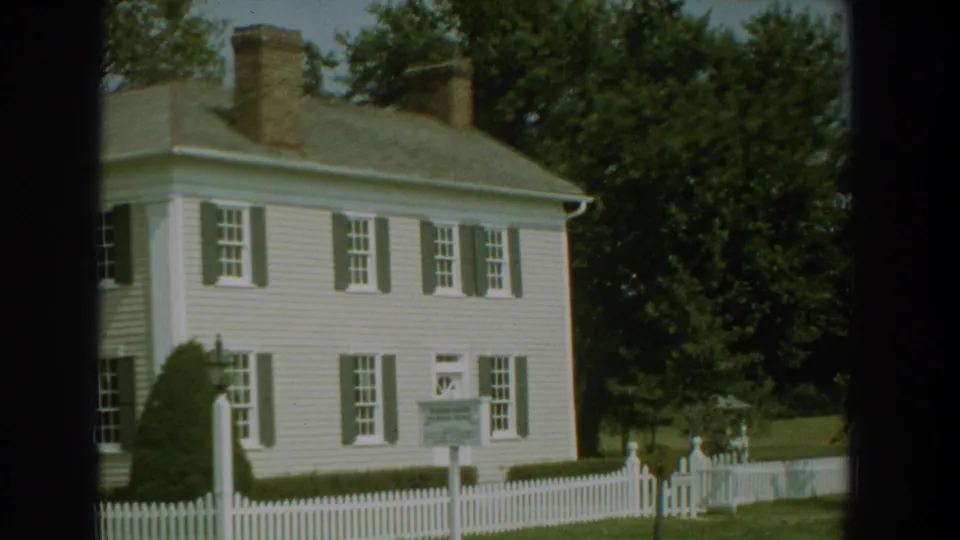 1959 Florida exterior view of a white clapboard-style house with shutters, chimneys, a picket fence, and landscaped greenery.