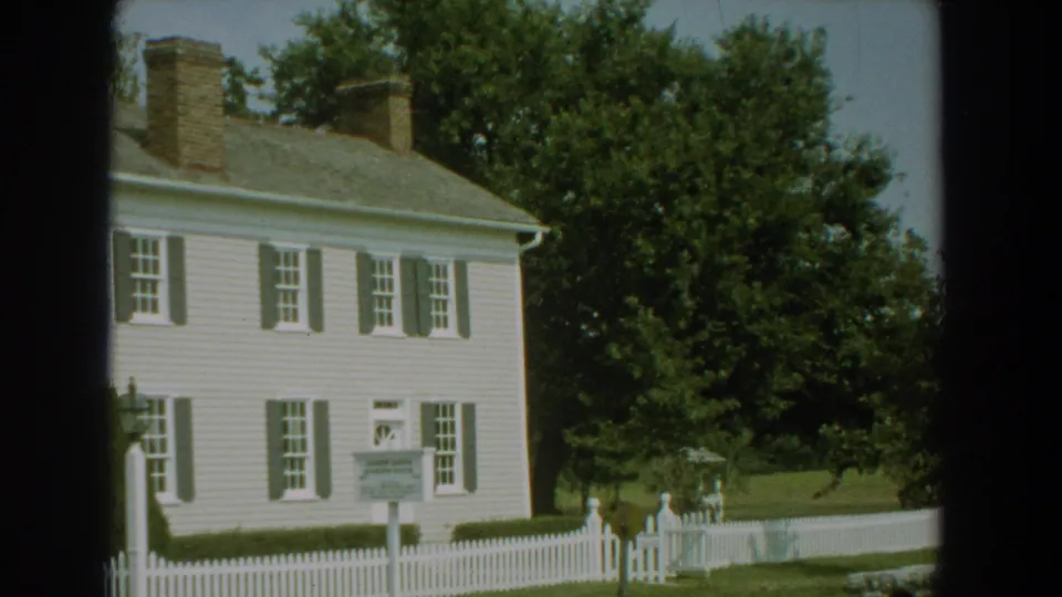 1959 Florida 8mm frame with the white house partly cropped, a signboard by the picket fence, green lawn, and a large tree.