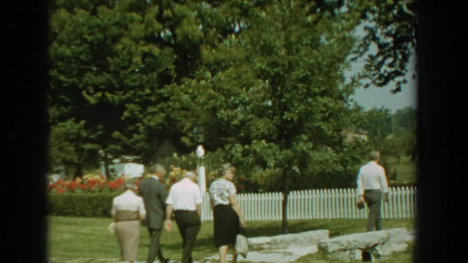1959 Florida 8mm frame showing senior visitors walking across lawn beside a white picket fence, trees, flowers, and a stone-lined path.