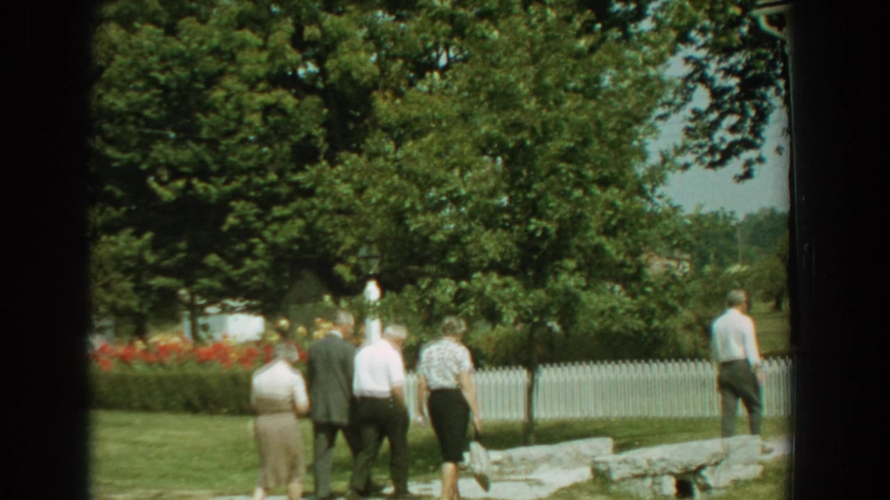 In 1959 Florida home-movie footage, adults walk through a sunny yard bordered by trees, flowers, a stone walkway, and a white picket fence.