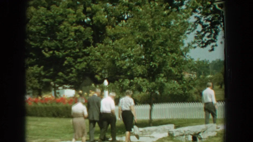 In 1959 Florida home-movie footage, adults walk through a sunny yard bordered by trees, flowers, a stone walkway, and a white picket fence.