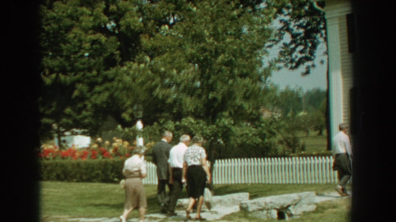 Group of adults continues along the lawn near a white fence and house edge.