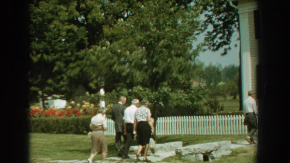 Adults in period clothing walk beside a white picket fence and garden plantings in a Florida residential yard from 1959 home-movie footage.