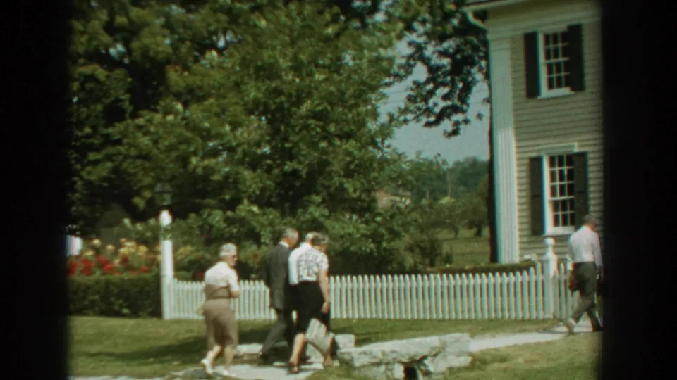Adults walk past a white fence and light-sided house, with trees and garden plantings visible in 1959 Florida home-movie footage.
