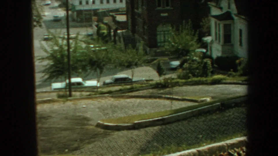 An elevated 1959 Florida home-movie view shows a quiet residential street with parked cars, houses, sidewalks, and shaded greenery.