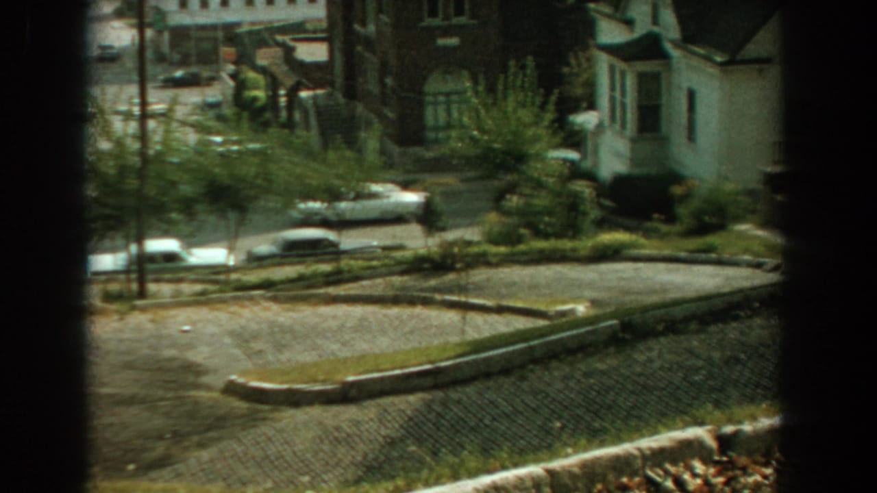 Elevated window view of a quiet neighborhood street with parked cars and low houses.